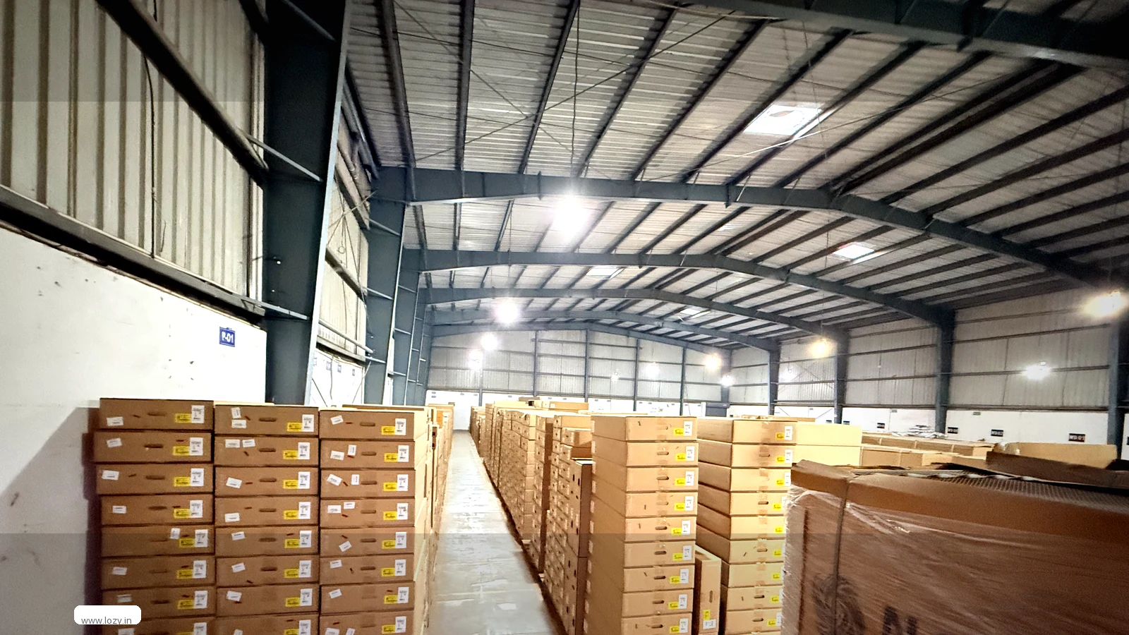 Block-stacked cartons in tall columns down a narrow aisle of a grey-clad warehouse, with a blue bay sign reading E01 on the wall and high-bay lights overhead
