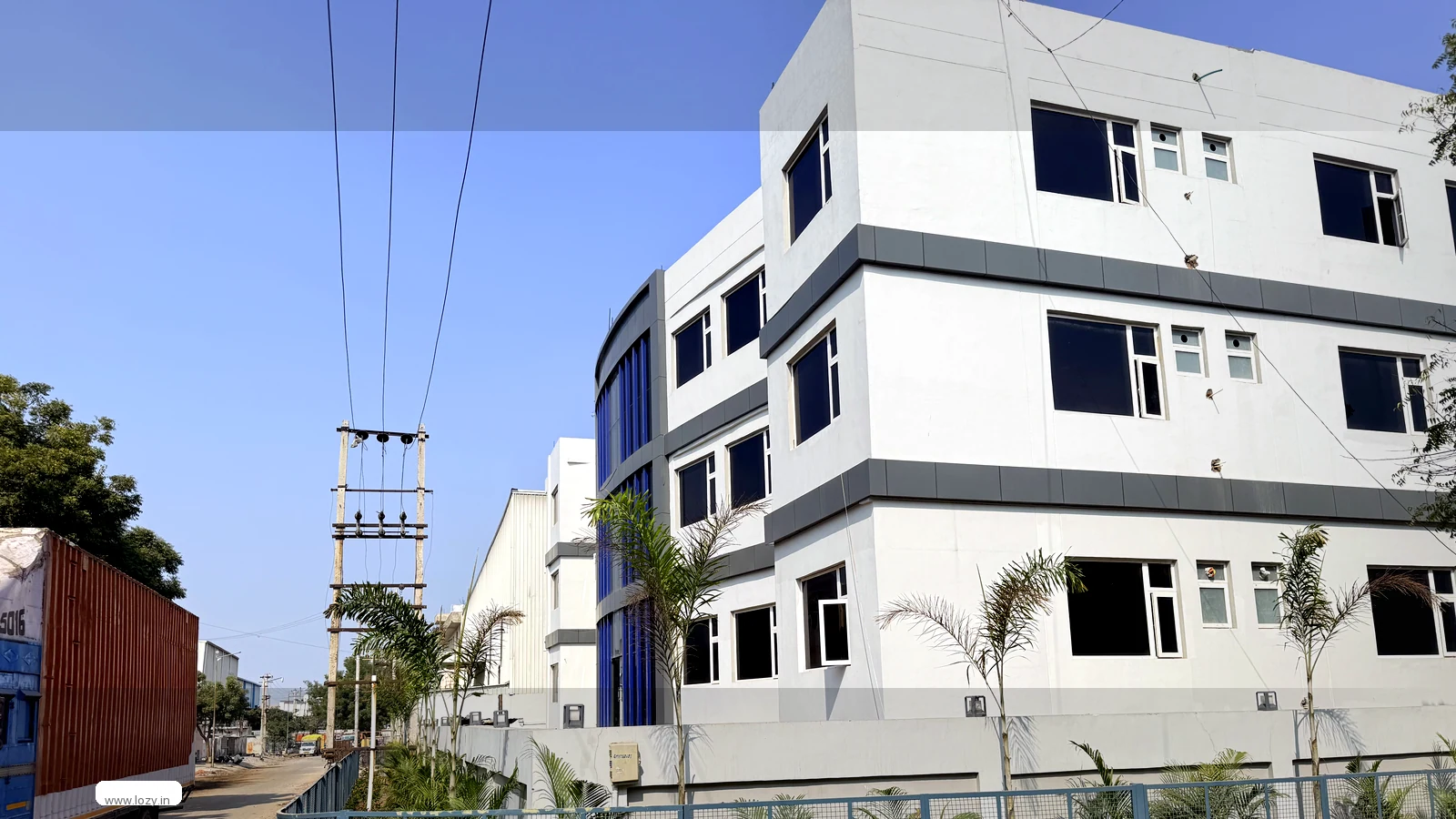 The Lozy warehouse building in Manesar Sector 8, a white multi-storey industrial block behind a blue mesh fence on the road into the township