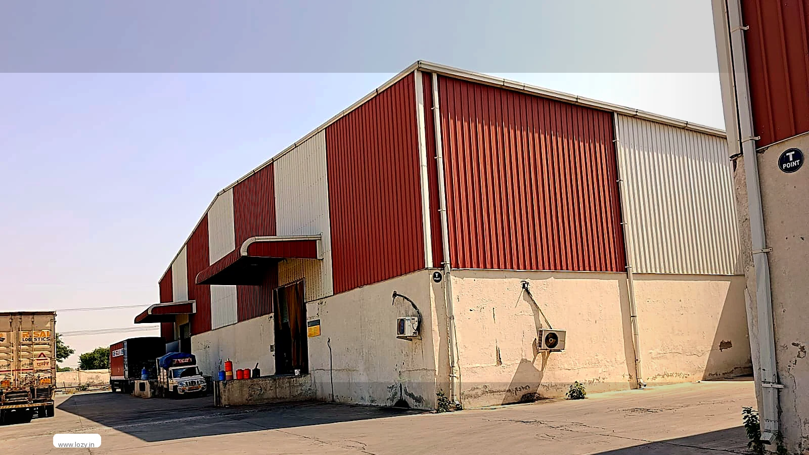 Single-storey Lozy warehouse at Farrukhnagar, Gurugram, with rust-red and off-white ribbed cladding, a loading doorway under a metal canopy and a concrete apron