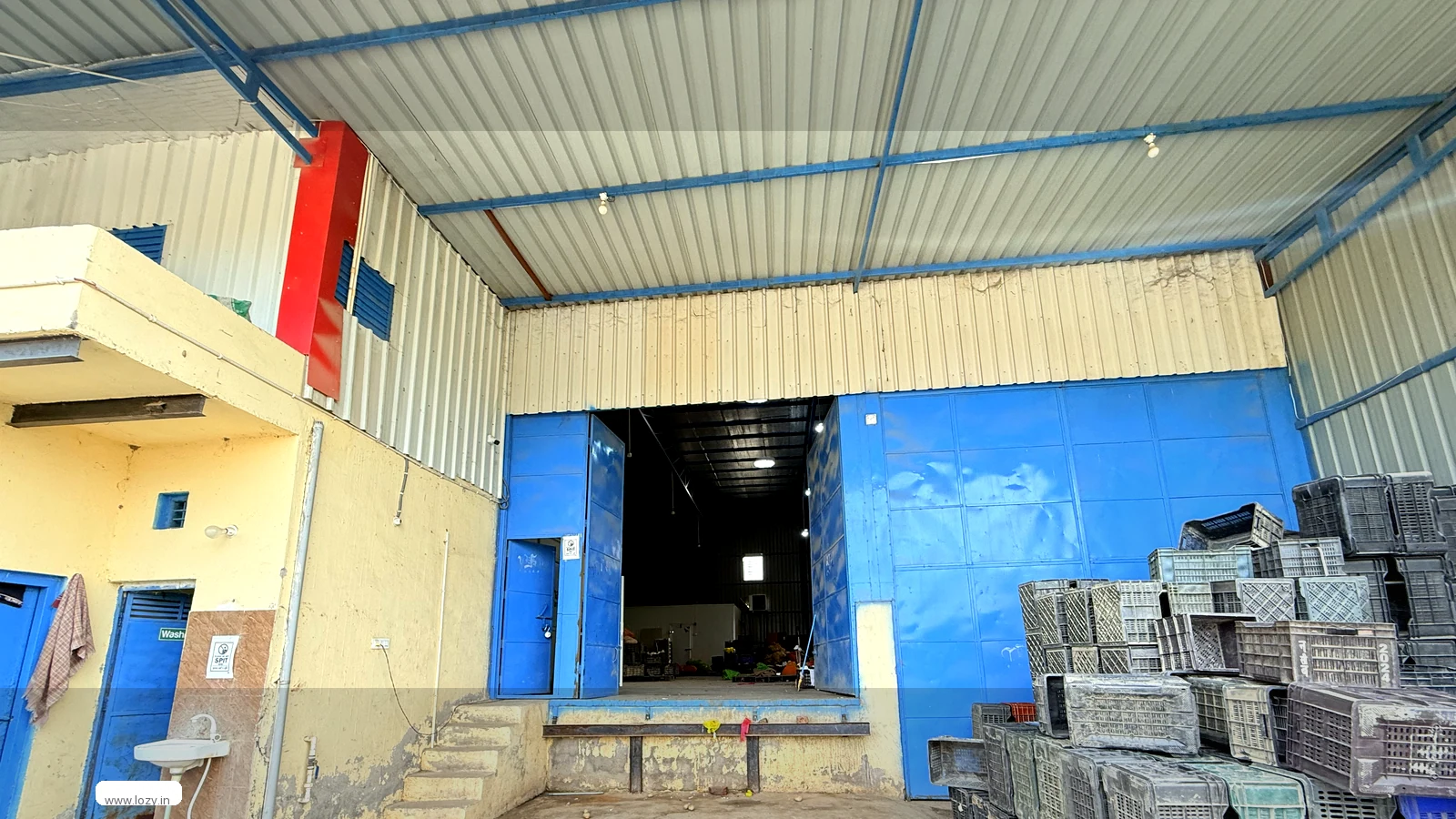 Inside a Lozy warehouse at Khirki Daula on the Gurugram side, with a blue steel portal frame, corrugated roof sheeting, a concrete slab floor and a folding door at the loading face
