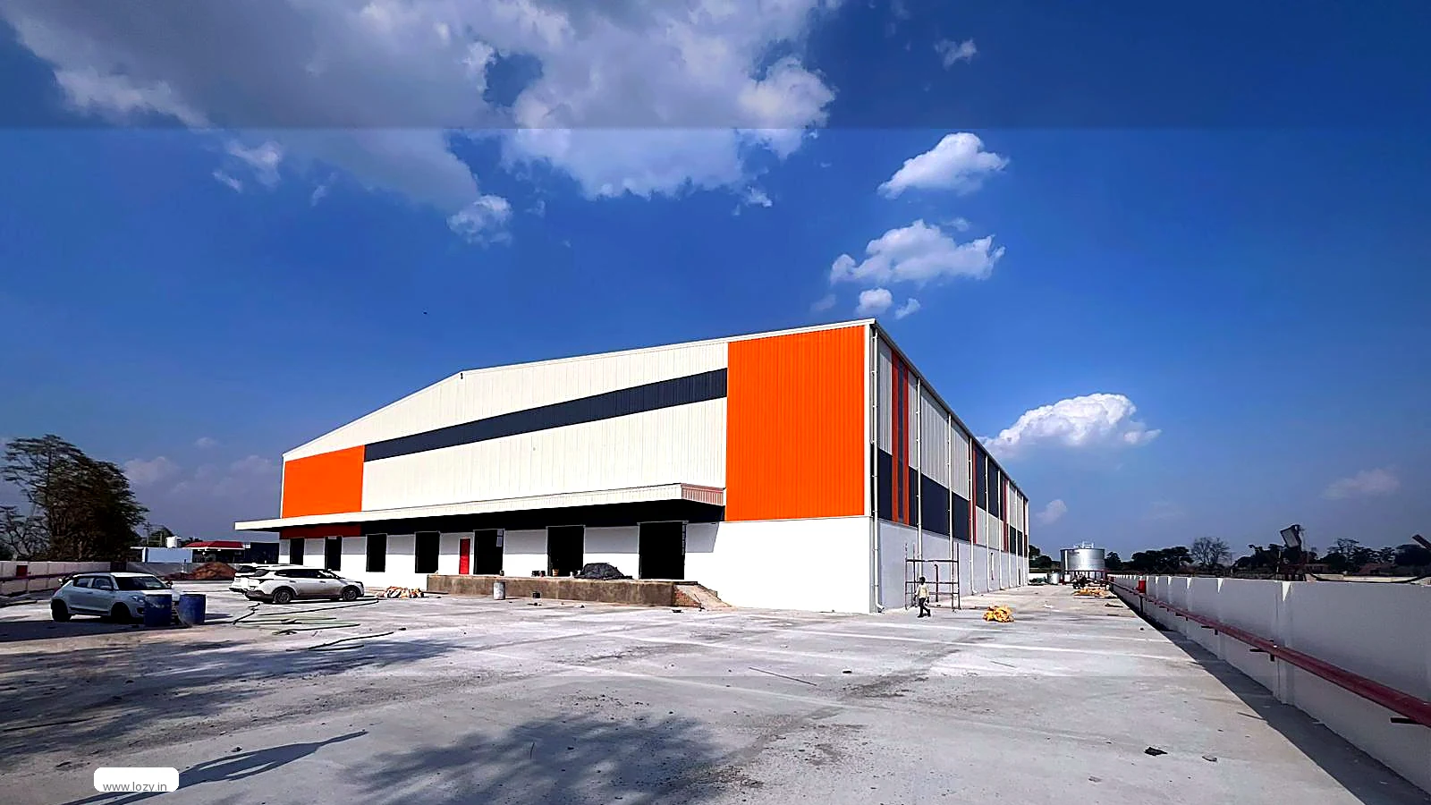 A single-storey clear-span warehouse shed with white cladding, an orange end wall and roller shutters opening onto a concrete apron and yard