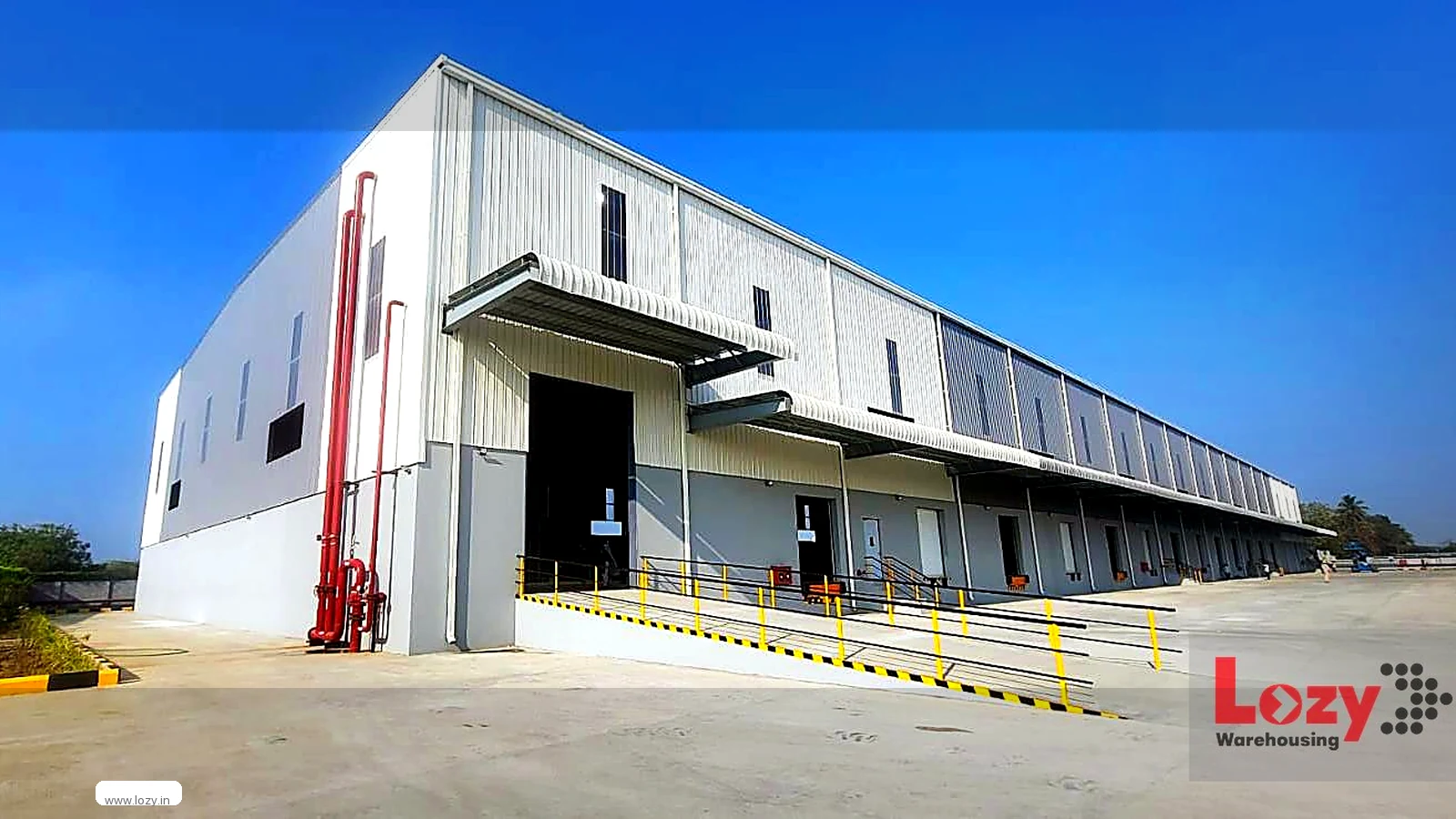 Exterior of a Lozy warehouse at Red Hills, Chennai — white corrugated cladding, a row of closed roller-shutter docks under individual canopies, a wide concrete ramp striped black and yellow, yellow safety railings and red fire standpipes at the corner