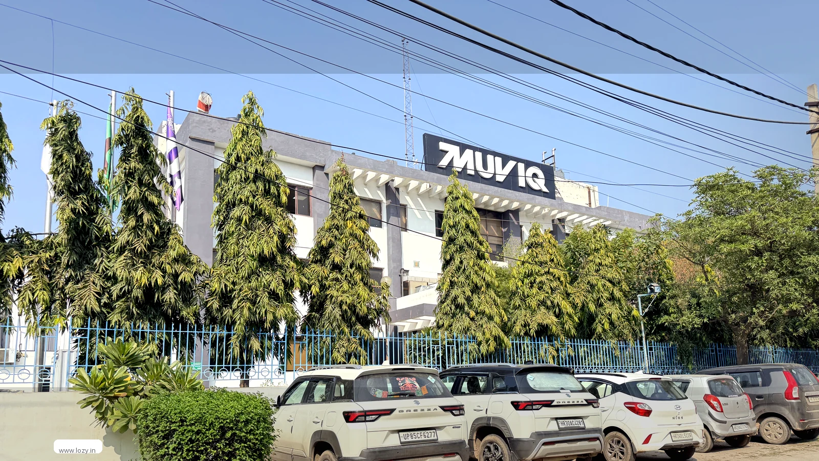 Warehouse property frontage seen from the road - a blue palisade fence, conifers along the boundary, a white banded building behind and three white SUVs on the paved forecourt