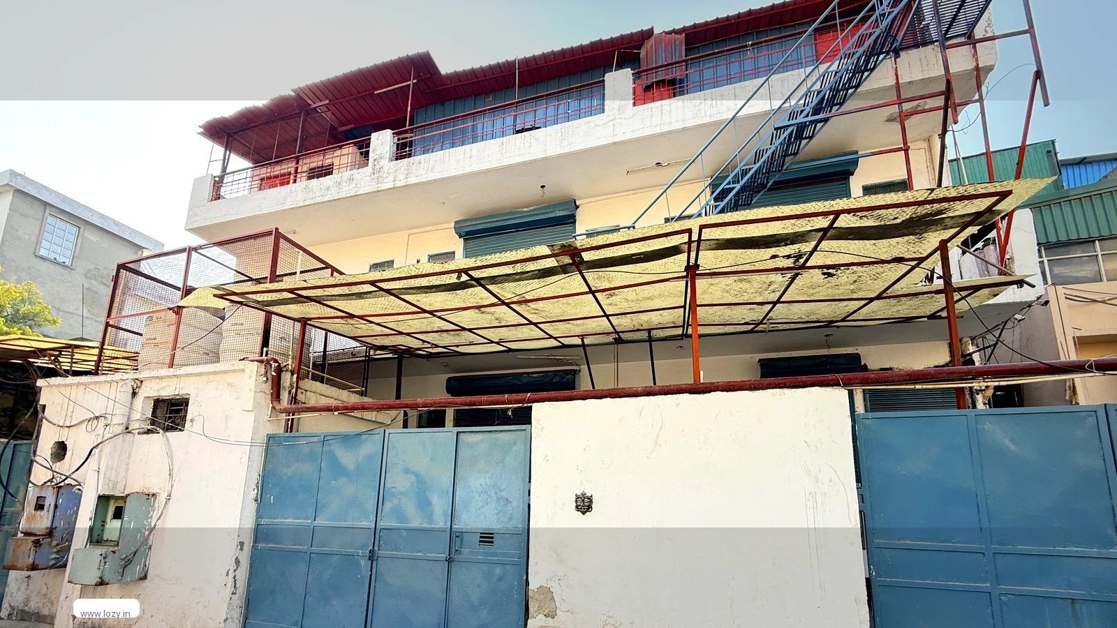 Industrial-area building in Faridabad seen from its compound, white walls with slate-blue metal gates and a concrete apron