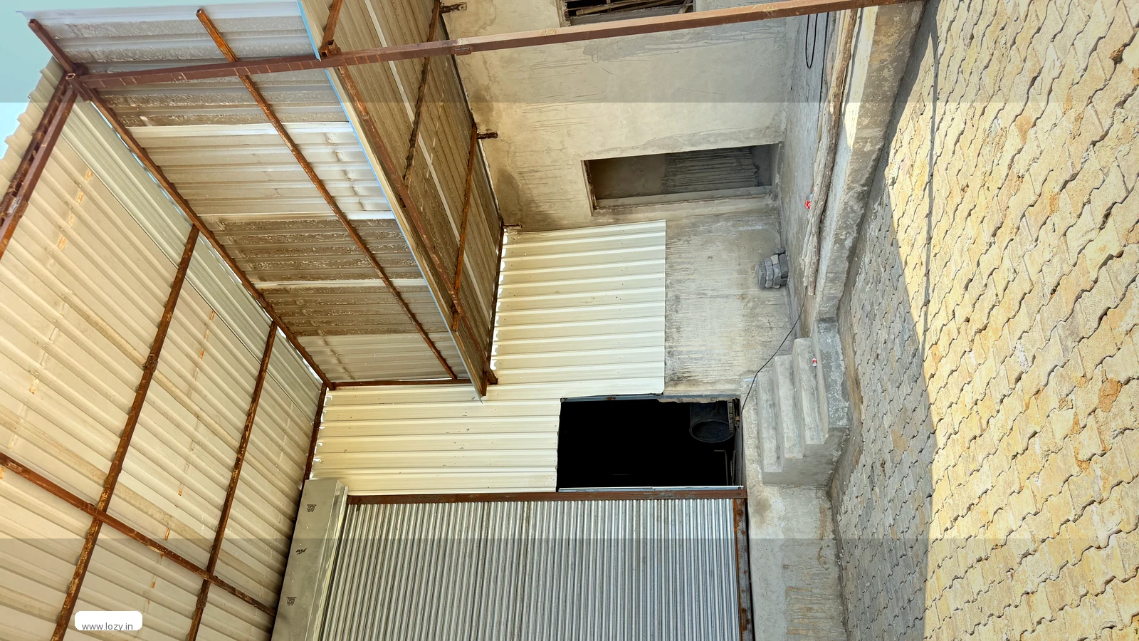 A closed corrugated roller shutter on a plain concrete warehouse block at Khirkai Daula, with a lean-to canopy on rusted steel framing and block paving across the front