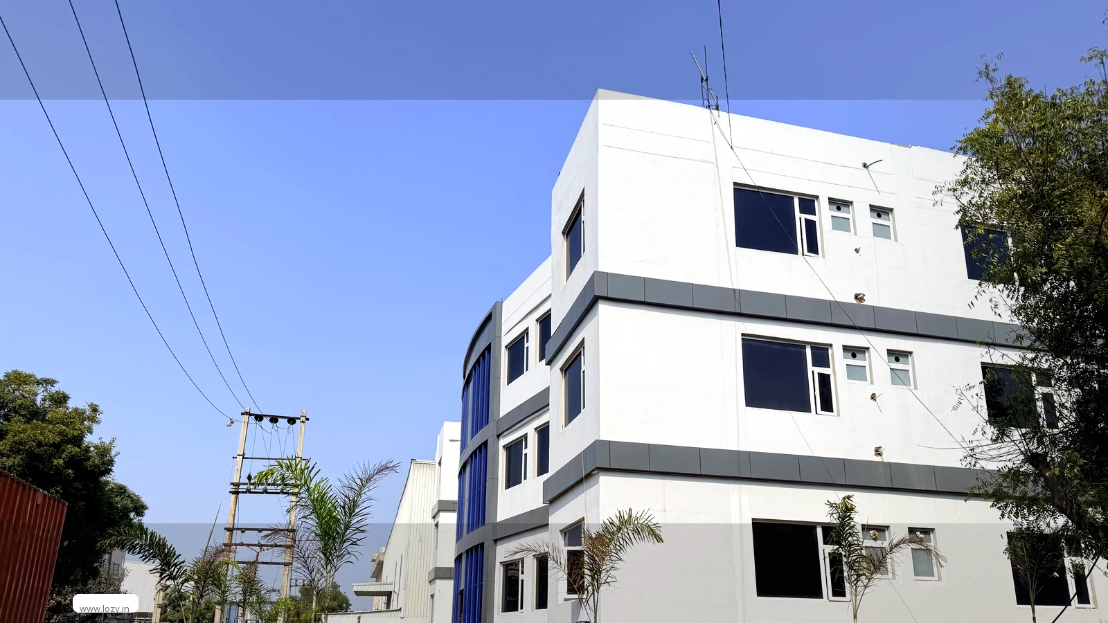Lozy facility at Manesar, Haryana - white multi-storey frontage with blue glazing beside a long metal-clad warehouse shed above the approach road